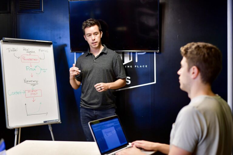 Mentor iz IT industrije objašnjava koncept razvoja softverskog projekta studentu-programeru tokom radionice, uz beleške i dijagrame na tabli.